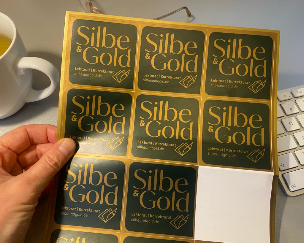 Bogen mit Silbe-und-Gold-Aufklebern, im Hintergrund Teetasse, Brille und Tastatur auf dem Schreibtisch