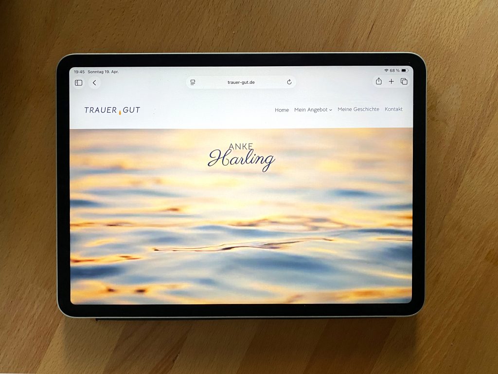 ipad auf heller Holzoberfläche. Abgebildet ist die Startseite von trauer-gut.de, mit dem Namenszug „Anje Harling" und einer Wasseroberfläche im Abendlicht